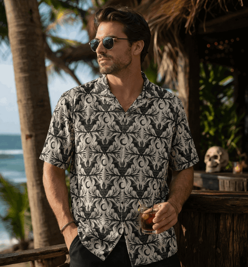 Man wearing a stylish bat crescent gothic button-up shirt by the beach, holding a drink with palm trees in the background.