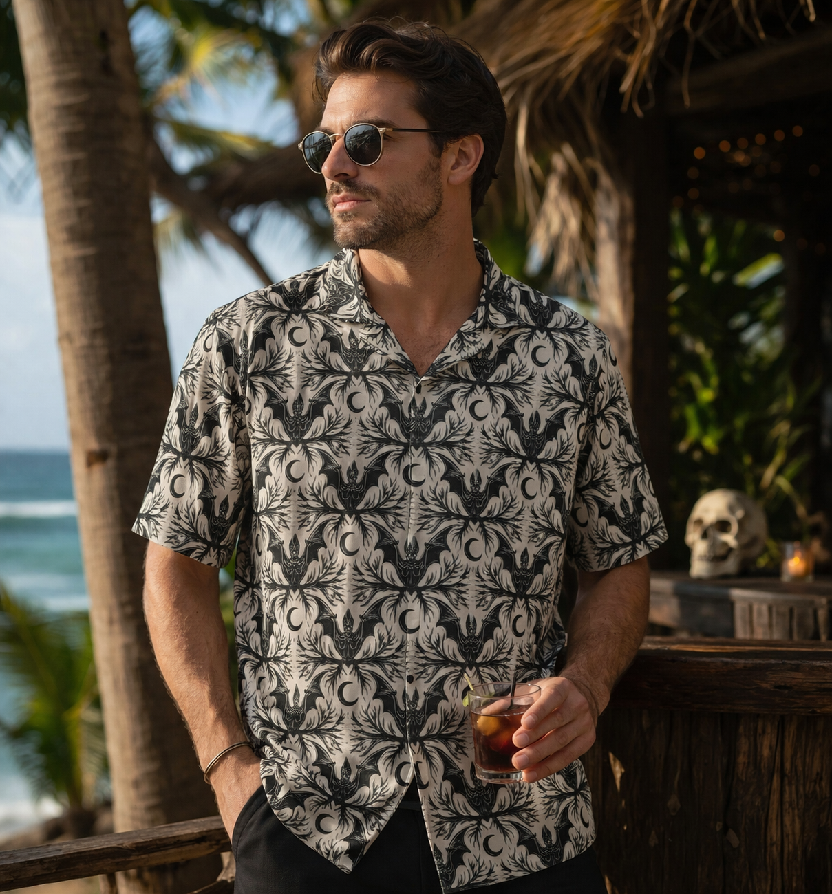 Man wearing a stylish bat crescent gothic button-up shirt by the beach, holding a drink with palm trees in the background.